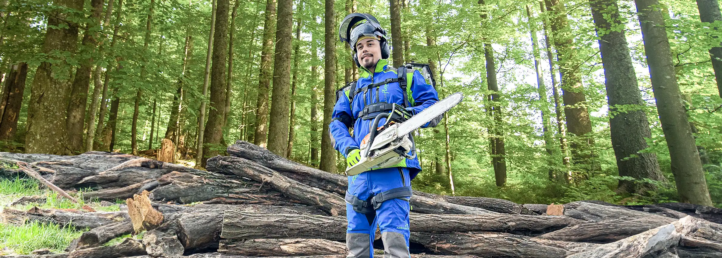 Mann mit blauer Forstwirtschaftskleidung und Exoskelett steht mit einer Motorsäge vor Bäumen im Wald