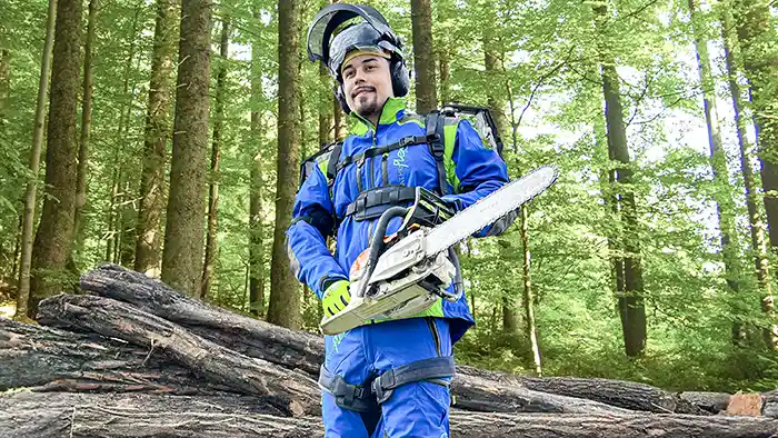 Mann mit blauer Forstwirtschaftskleidung und Exoskelett steht mit einer Motorsäge vor Bäumen im Wald