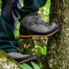 Graue Haix Ramble GTX-Schuhe mit orangen Akzenten an den Füßen einer Person, die auf einem Baumstamm im Wald steht.