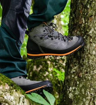 Graue Haix Ramble GTX-Schuhe mit orangen Akzenten an den Füßen einer Person, die auf einem Baumstamm im Wald steht.