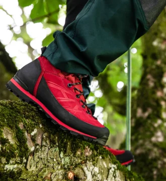 Ein roter Haix Ramble GTX-Schuh an einem Fuß, der auf einem moosbewachsenen Ast steht, umgeben von grünem Laub im Wald.