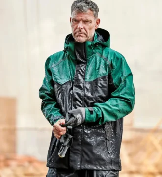 Mann trägt die grün-schwarze Elka Regenjacke im strömenden Regen – wetterfeste Arbeitsjacke mit Kapuze und hohem Kragen im Einsatz auf der Baustelle